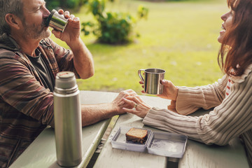 Fototapeta premium Happy middle-aged couple in love sitting on the benches with table in park, smile and drink tea or coffee from thermos.