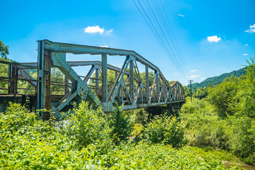 Railway bridge over river in Polish mountain. Heavy rusty steel old industrial train overpass in natural scenic landscape, blue summer sky.