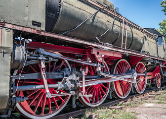 Naklejka premium Huge vintage steam locomotive, red painted steel wheel detail close up. Coal-powered steam train stands on a siding. Classic gigantic heavy railway machinery. Side view of power parts of machine.