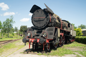 Obraz premium Huge vintage steam locomotive, red painted steel wheel detail close up. Coal-powered steam train stands on a siding. Classic gigantic heavy railway machinery. Side view of power parts of machine.