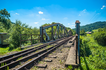 Railway bridge over river in Polish mountain. Heavy rusty steel old industrial train overpass in natural scenic landscape, blue summer sky.