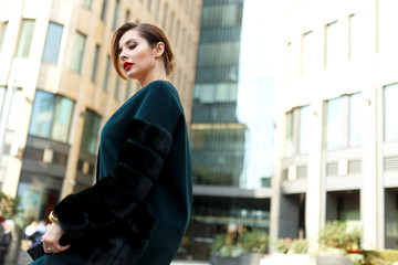 Side virew of a beautiful stylish woman walking down the street. Fashionable girl wearing dark green fur coat and clutch.