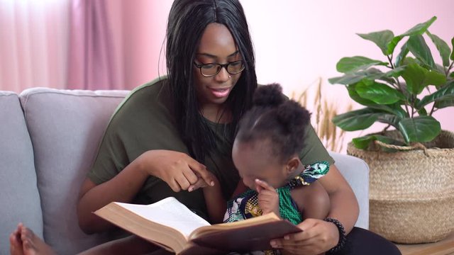 african american mother reading a book to her little daughter on couch at home . black girl lying on her mom study at home on sofa together . quarantine Home School