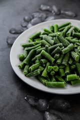 a white plate with frozen green string beans covered with melting ice and hoarfrost. frozen green vegetables product. winter food preservation. cold from the freezer. dark vertical selective focus