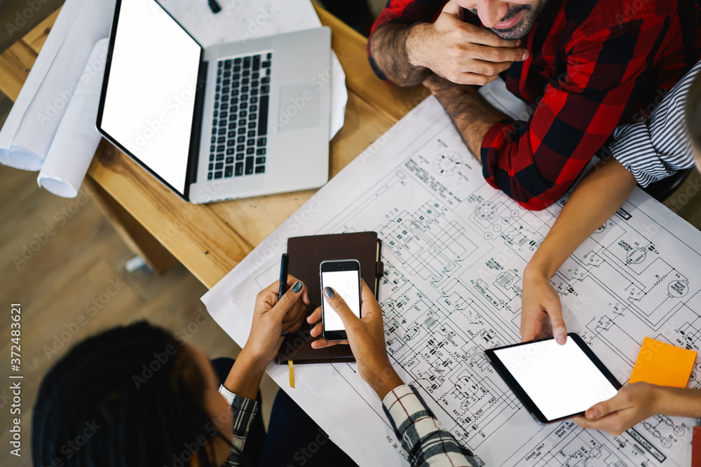 Poster cropped top view of hipster guys sitting at sketches and blueprints using modern smartphones and lap