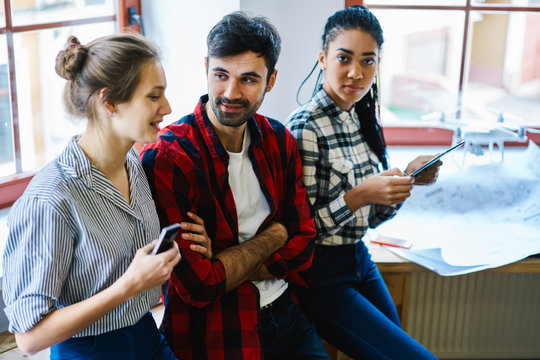 Young smiling woman telling funny story about misunderstanding to male and female colleagues dressed in casual outfit during break from drawing building sketches on work sitting in coworking space