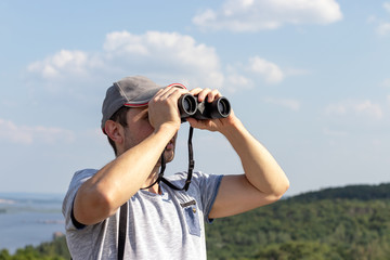 A man looks through binoculars against a scenic view of a wide river on a hill on a sunny day