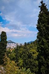 Fototapeta na wymiar View of Lycabettus hill from Areopagus hill with rich foliage and high cedar or cypress trees. Athens, Greece. Summer day and blue sky with clouds