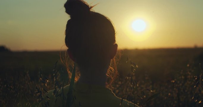 Back portrait of girl with bun praying alone outdoors looking at golden setting sun. Unrecognizable woman standing in evening field holding joined hands handheld device. Meditation concentration