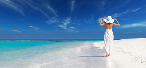 elegant woman in white dress holds sun hat and walks on idyllic beach of maldives, relax in a tropical island - Powered by Adobe