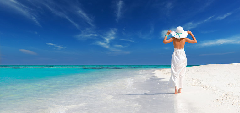 elegant woman in white dress holds sun hat and walks on idyllic beach of maldives, relax in a tropical island