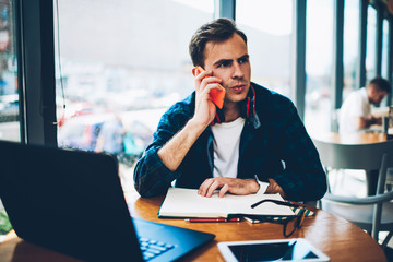 Young good looking male person communicating via smartphone connected to wireless internet while sitting in coffee shop.Professional freelancer having mobile conversation with colleague indoors