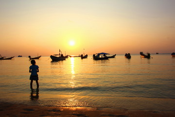 girl is standing alone on beach and looking to the group of  long tail boats parking on coast with sun aet background.