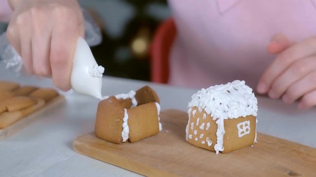 Woman is making gingerbread house glues parts with sugar sweet icing, hands closeup. Cooking and decorating homemade gingerbread house for Christmas holidays. New Year family traditions.