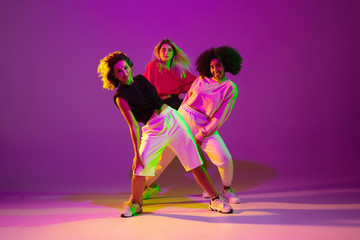 Flexible. Sportive girls dancing hip-hop in stylish clothes on purple-pink background at dance hall...