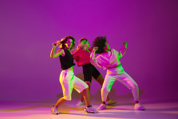 Flexible. Sportive girls dancing hip-hop in stylish clothes on purple-pink background at dance hall...