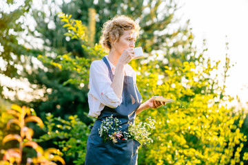 Beautiful portrait of smiling middle-aged woman with wild flowers in pocket, holding secateurs in garden. She is absolutely happy. Slow living, gardening hobby concept