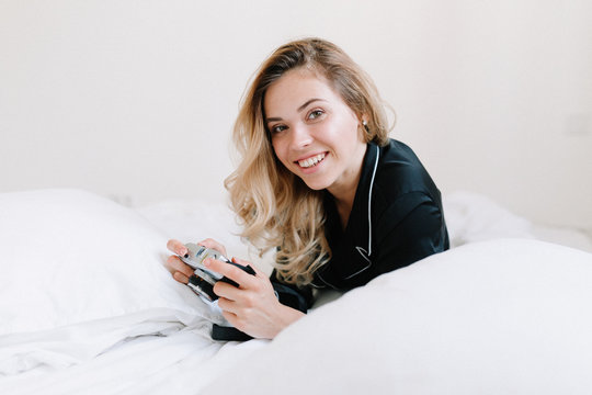Indoor portrait of european lovely girl with blonde hair lying on the white bed and holding retro camera. Morning at home