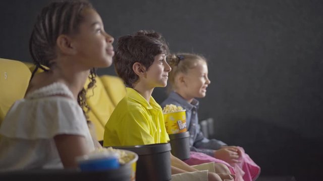 Side view of cute Middle Eastern boy watching cartoon in cinema with friends. Multiethnic children enjoying film in movie theatre. Carefree kids sitting on yellow chairs and laughing.