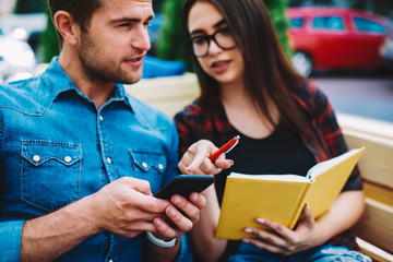 Cropped image of two students communicating about received sms message on modern telephone.Hipster girl in eyeglasses and book in hands pointing to friend right direction on mobile navigator online