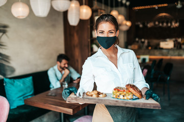 Beautiful young waitress with face protective mask serving delicious burger to middle age male customer. Corona virus and small business is open for work concept. - obrazy, fototapety, plakaty