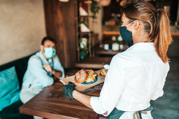 Beautiful young waitress with face protective mask serving delicious burger to middle age male...