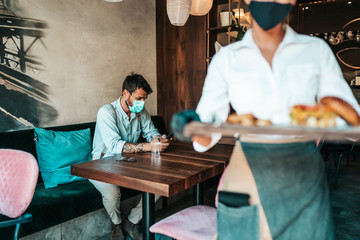 Beautiful young waitress with face protective mask serving delicious burger to middle age male customer. Corona virus and small business is open for work concept.