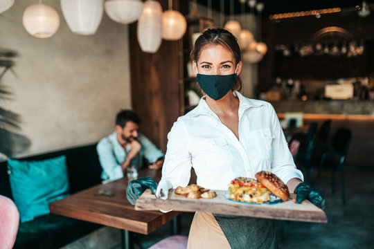 Beautiful Young Waitress With Face Protective Mask Serving Delicious Burger To Middle Age Male Customer. Corona Virus And Small Business Is Open For Work Concept.