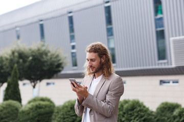 Portrait of handsome curly businessman in casual wear holding smartphone and smiling.Successful manager using mobile phone apps,texting message,browsing internet,looking at phone near business center.