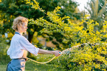 Housewife working in garden. Happy blond woman gardener watering garden with hose. Hobby, leisure...