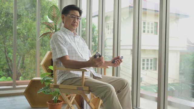 Asian elderly man sitting on chair and using hands grip exercise for care and treatment health in living room at alone home. Healthcare and fitness concept of senior or oldman.
