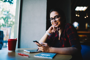 Half length portrait of smiling gorgeous female blogger looking at camera while sharing news and ideas with followers on personal website using modern telephone connected to 4G internet indoors