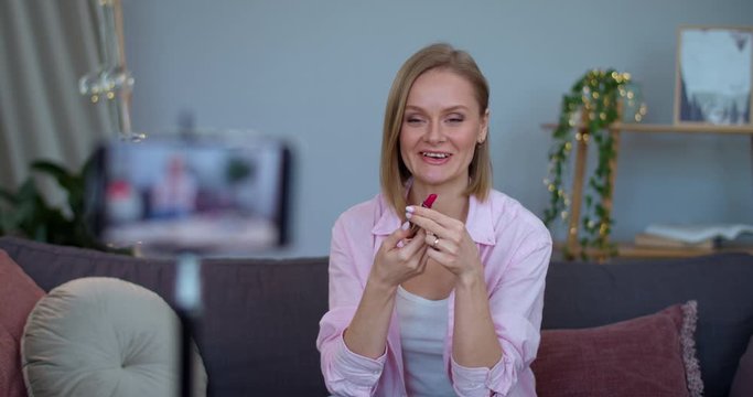 Smiling woman open and showing lipstick to camera while sitting on sofa in front of smartphone. Female vloger talking while recording make up review. Concept of beauty and vlogging.