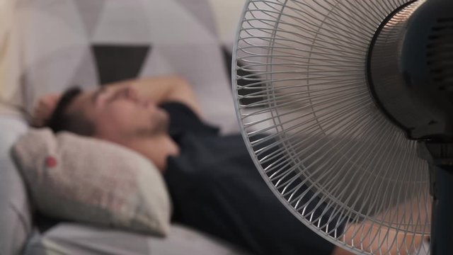 Hot young man lying on the living room couch with the fan on. Concept of heat in summer