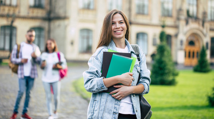 Beautiful young student girl with a backpack and books is looking on camera on group of her friends background in university