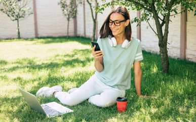 Young modern beautiful happy confident woman in headphones with a smartphone is working on a laptop while sitting on the lawn.