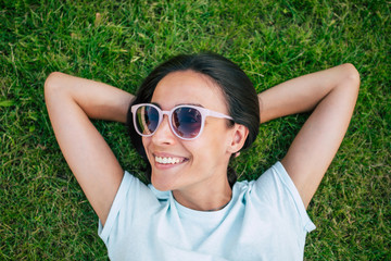 Happy beautiful hipster woman in cool sunglasses is having fun while she is lying on the lawn and looking on camera