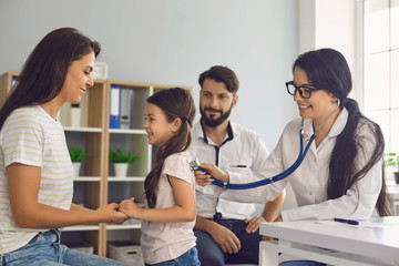 Fototapeta premium The family doctor listens with a stethoscope to the child with the parents sitting at the table in the clinic.