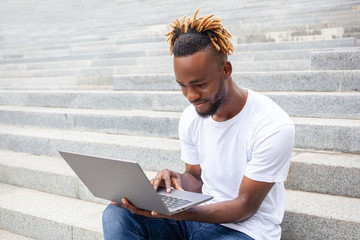 Freelance, technology, and education concept. Young concentrated African American guy working on laptop outdoors , sitting on the stairs in park. Empty copy space.