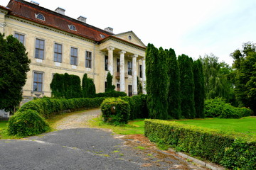 Fototapeta na wymiar View of an old abandoned and slightly ruined mansion or palace with many windows, arch based entrance, and a slanted red roof surrounded with beautidul gardens, hedges, and tall trees seen in Poland