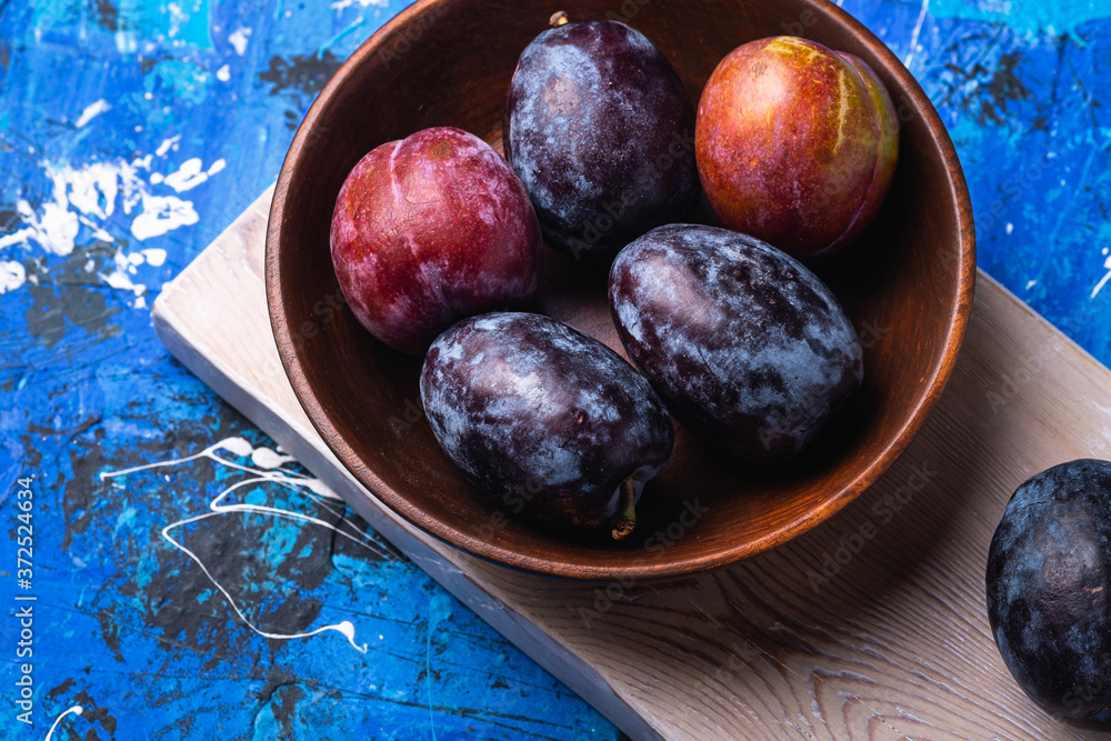 Wall mural fresh sweet plum fruits in brown wooden bowl on old cutting board, blue abstract background, angle v