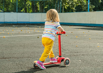 Little girl with blond wavy hair in sportswear with a kick scooter on a fenced sports ground. Photo...
