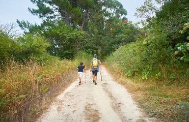 Fototapeta premium Mother with her son walking with backpacks and walking sticks along a path in the field full of trees