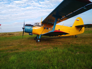 An old yellow biplane with blue wings and a running engine drives through a field of green grass in...