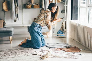 Brunette woman holds smartphone and makes photos of clothes to sell after childbirth via internet. Young woman consults with a designer stylist at home about her wardrobe. - obrazy, fototapety, plakaty