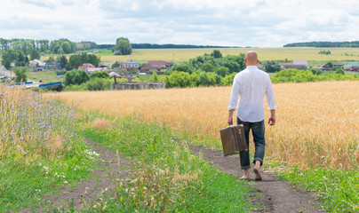 A brutal young man in a white shirt goes barefoot on the way home holding an old large suitcase. A man returns after traveling to his native land.