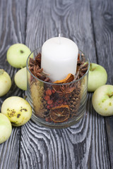 A candle in a glass candlestick. The candlestick is filled with dried cones, spices and dried fruits. Apples lie on the table surface nearby.