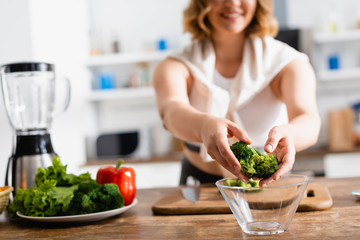 cropped view of woman putting broccoli in bowl near vegetables