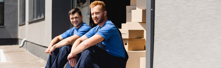 Obraz premium Panoramic shot of movers looking at camera while sitting in warehouse outdoors