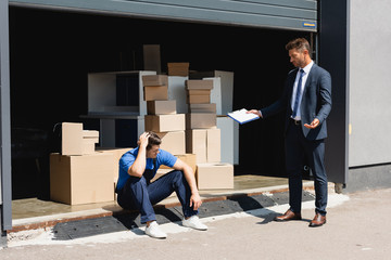 Businessman in suit holding clipboard and pointing with hand near sad loader in warehouse